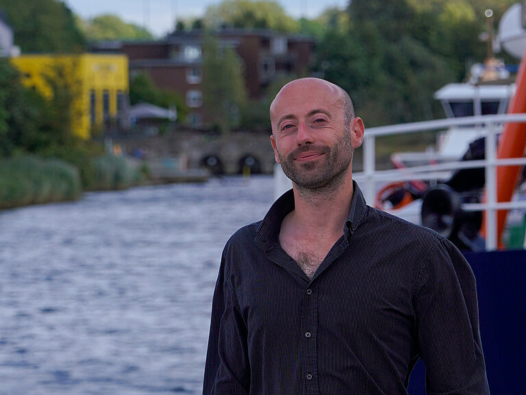 Ein junger Mann mit kurzem Bart und Glatze steht vor einem Fluss und lächelt in die Kamera. er trägt ein schwarzes Hemd.