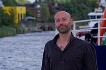 A young man with a short beard and a bald head is standing by a river, smiling at the camera. He is wearing a black shirt.
