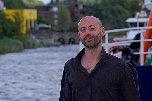 Ein junger Mann mit kurzem Bart und Glatze steht vor einem Fluss und lächelt in die Kamera. er trägt ein schwarzes Hemd.
