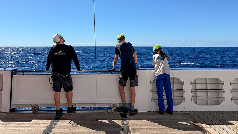Three people are standing on board a research vessel, watching as a piece of equipment is lowered into the water