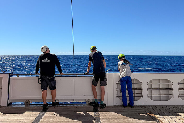 Drei Personen stehen an Bord eines Forschungsschiffes und schauen dabei zu, wie ein Gerät ins Wasser gelassen wird 
