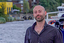 Ein junger Mann mit kurzem Bart und Glatze steht vor einem Fluss und lächelt in die Kamera. er trägt ein schwarzes Hemd.
