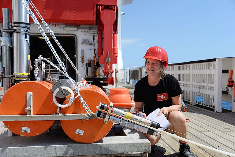  Woman with red hard hat and clipboard in hand squats on a ship deck in front of a device.