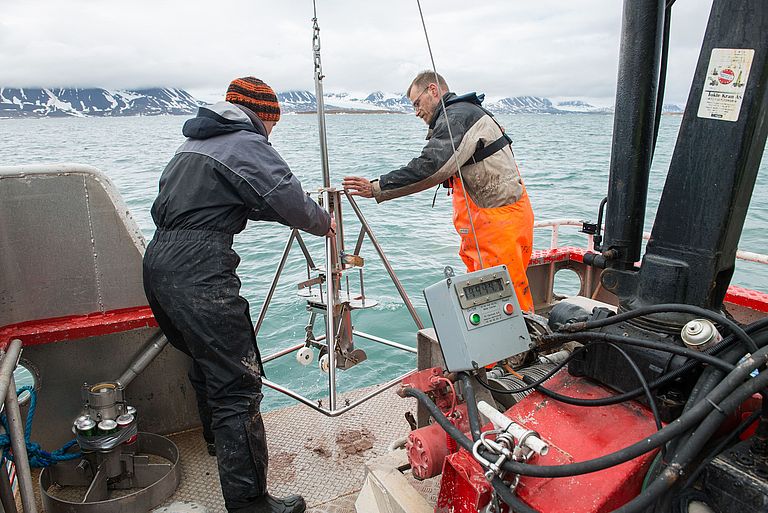 A man and a woman in work clothes stand at the stern of a ship and hold a scientific instrument in their hands. Glaciers can be seen in the background.  Photo: Bo Barker Jørgensen 