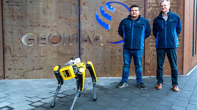 Ein gelber Roboterhund steht vor einem Gebäude mit dem Schriftzug "GEOMAR", dahinter zwei Männer in blauen Jacken