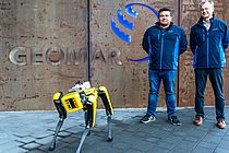 A yellow robot dog stands in front of a building with the word "GEOMAR" on it, with two men in blue jackets behind it.