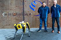 Ein gelber Roboterhund steht vor einem Gebäude mit dem Schriftzug "GEOMAR", dahinter zwei Männer in blauen Jacken