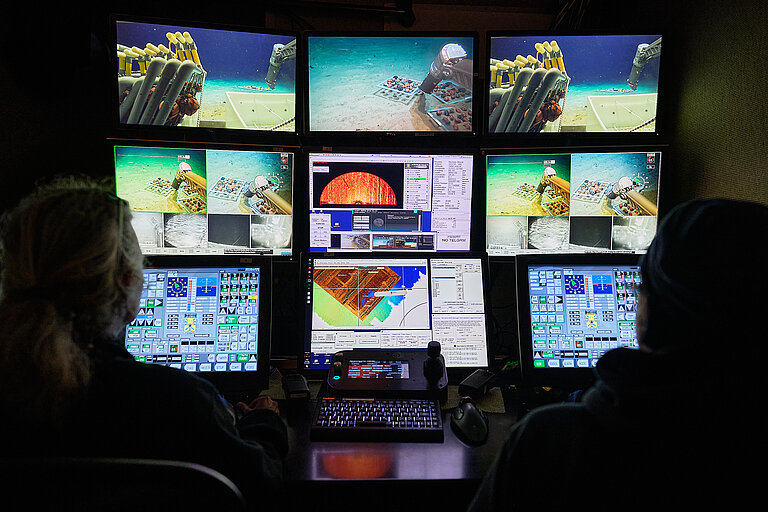 Nine monitors glow in the dark; two people are sitting in front of them