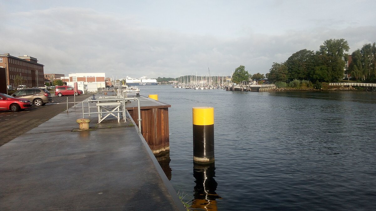 the kay in front of the Geomar building and the Schwntine river can be seen. Two large black pillars reach out of the water that mark and protect the area where the pipeline reaches the institute. 