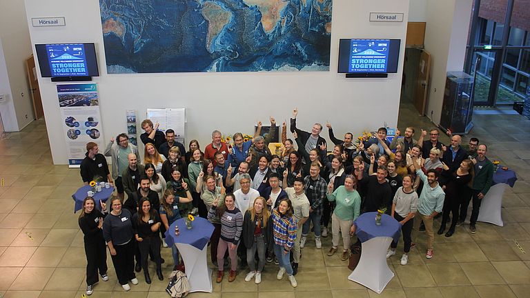 A group of people are standing in a foyer. Some of them point to a screen with the words "Stronger together".