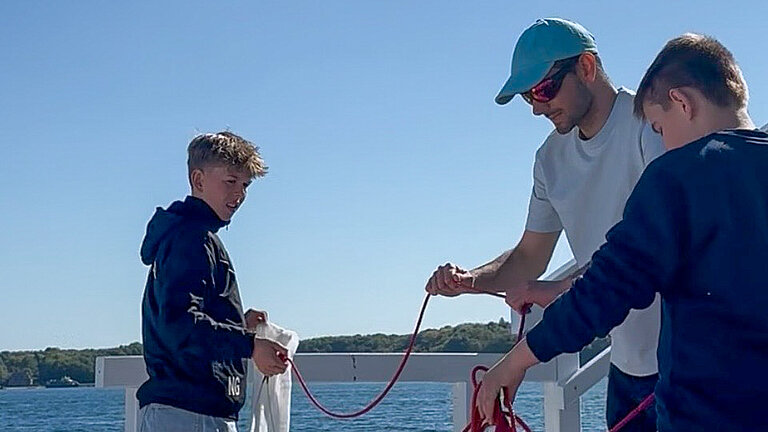 A young man wearing a light blue cap and sunglasses is standing by the sea with two lads in hoodies; they are holding lines in their hands.