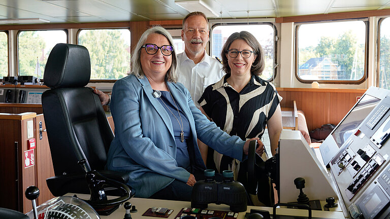 On the bridge of a ship: a woman is sitting in the helmsman’s seat, whilst the captain and another woman are standing behind her.