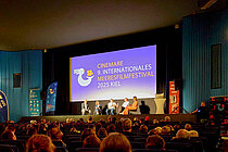A packed movie theater. A panel discussion is taking place on stage. 